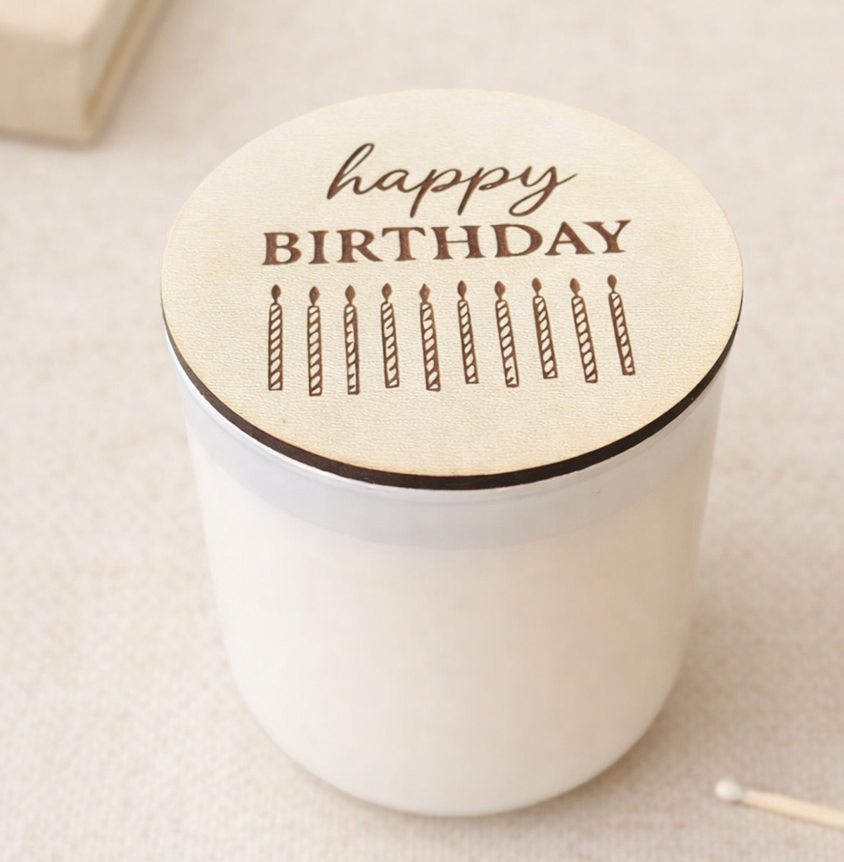 White candle with a wooden lid featuring 'happy birthday' and candles on a light background